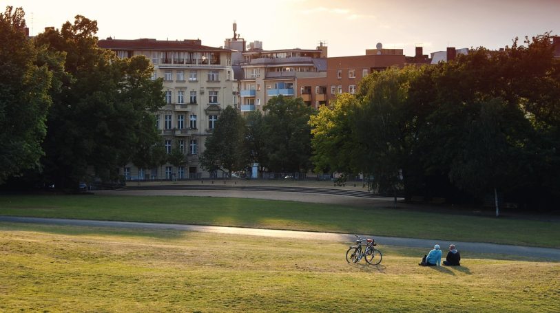 Grüne Wiese mit Menschen und Fahrrädern, im Hintergrund Stadtgebäude bei Sonnenuntergang.
